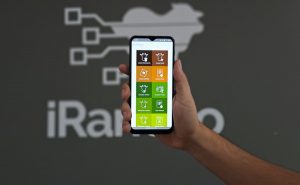Na foto, temos uma mão segurando um celular com a tela inicial do aplicativo iRancho. Ao fundo, observamos uma parede cinza com a logo do iRanho. Foto ilustra matéria de coleta de dados no curral. 