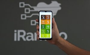 Na foto, temos uma mão segurando um celular com a tela inicial do aplicativo iRancho. Ao fundo, observamos uma parede cinza com a logo do iRanho. Foto ilustra matéria de coleta de dados no curral.
